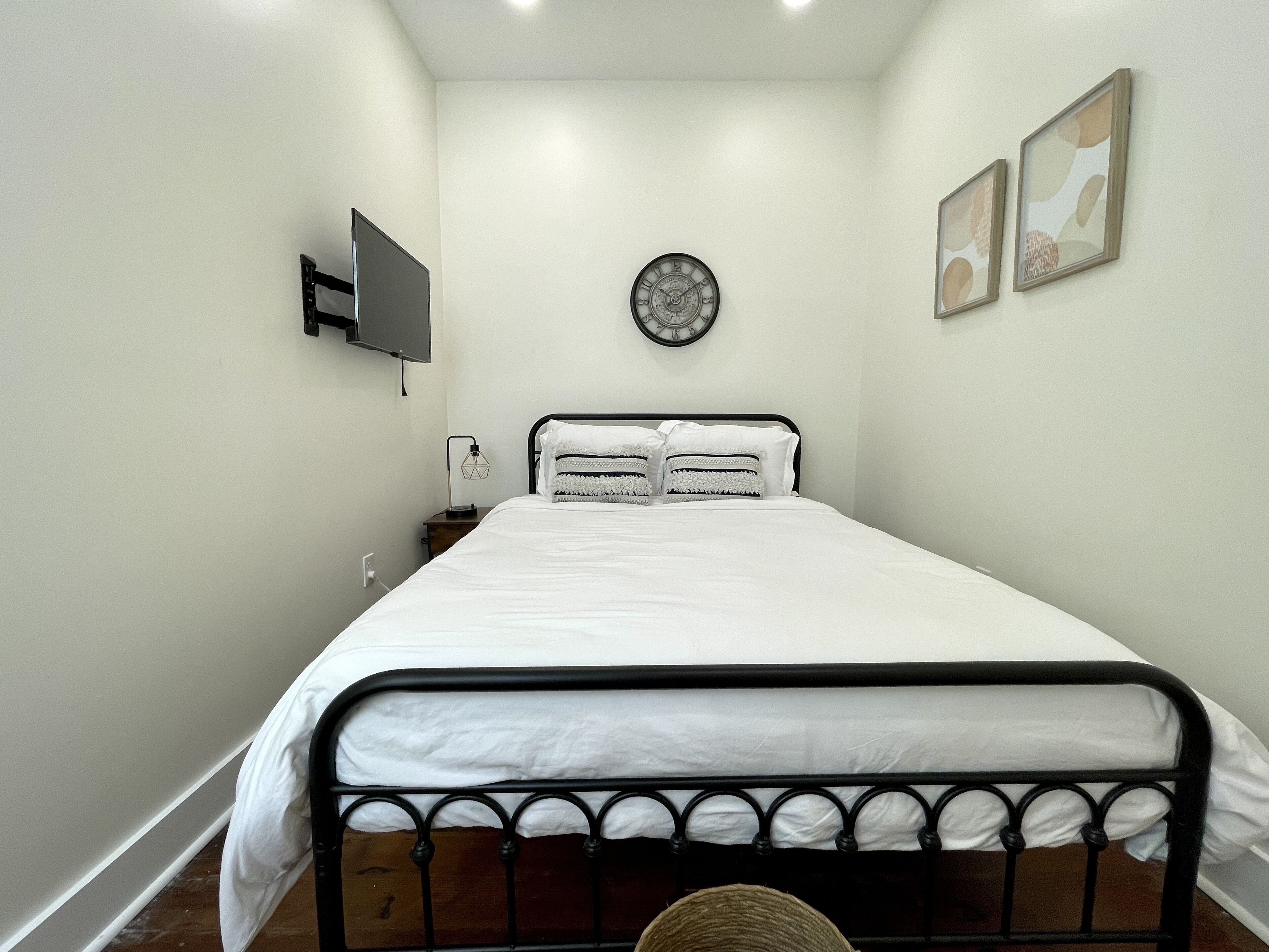 Un dormitorio ordenado con una cama, un reloj de pared, un televisor de pantalla plana y dos cuadros en la pared.