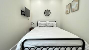 Un dormitorio ordenado con una cama, un reloj de pared, un televisor de pantalla plana y dos cuadros en la pared.