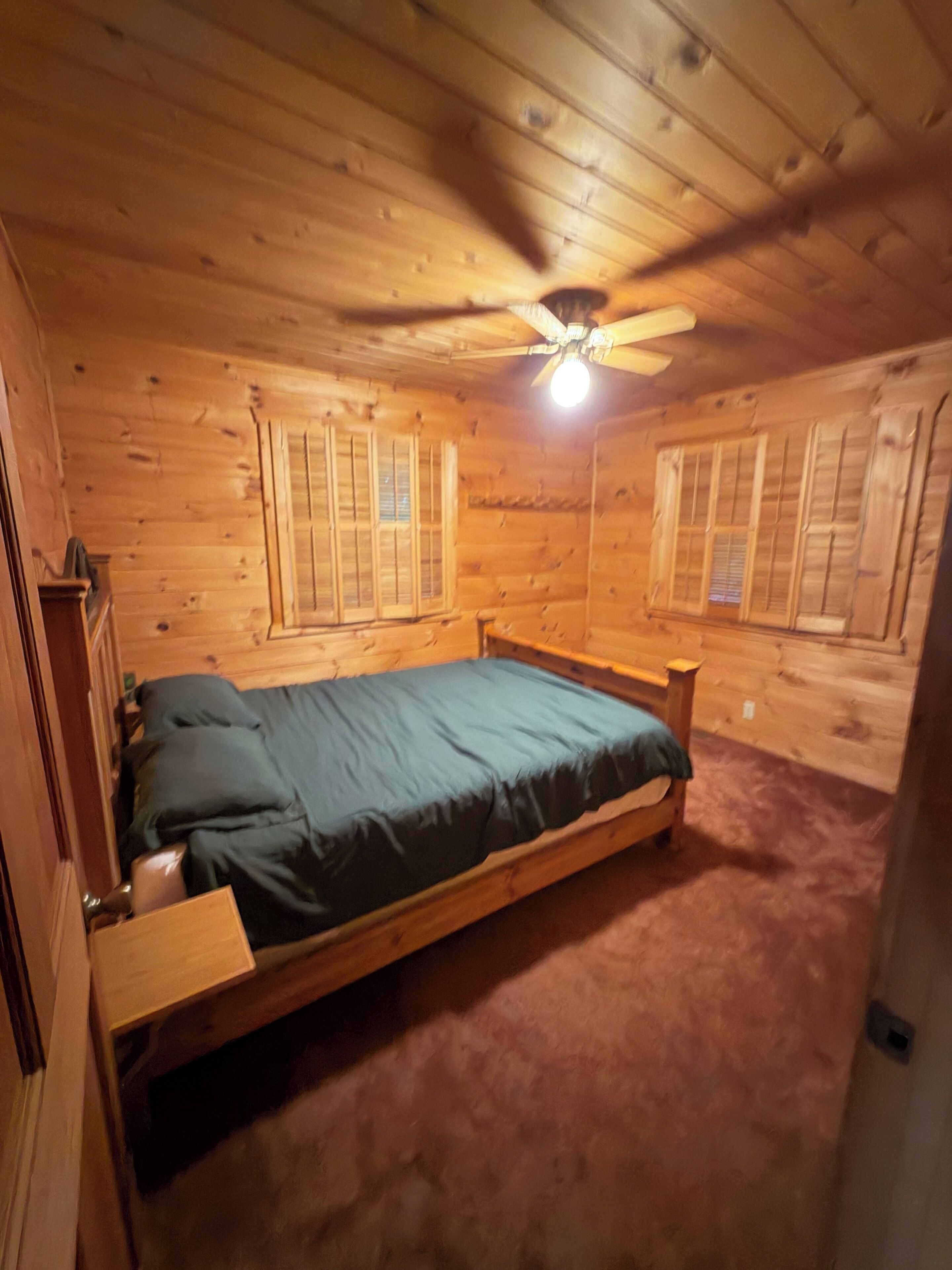 Une chambre dans une cabane en bois, avec un lit, un ventilateur de plafond et deux fenêtres équipées de volets.