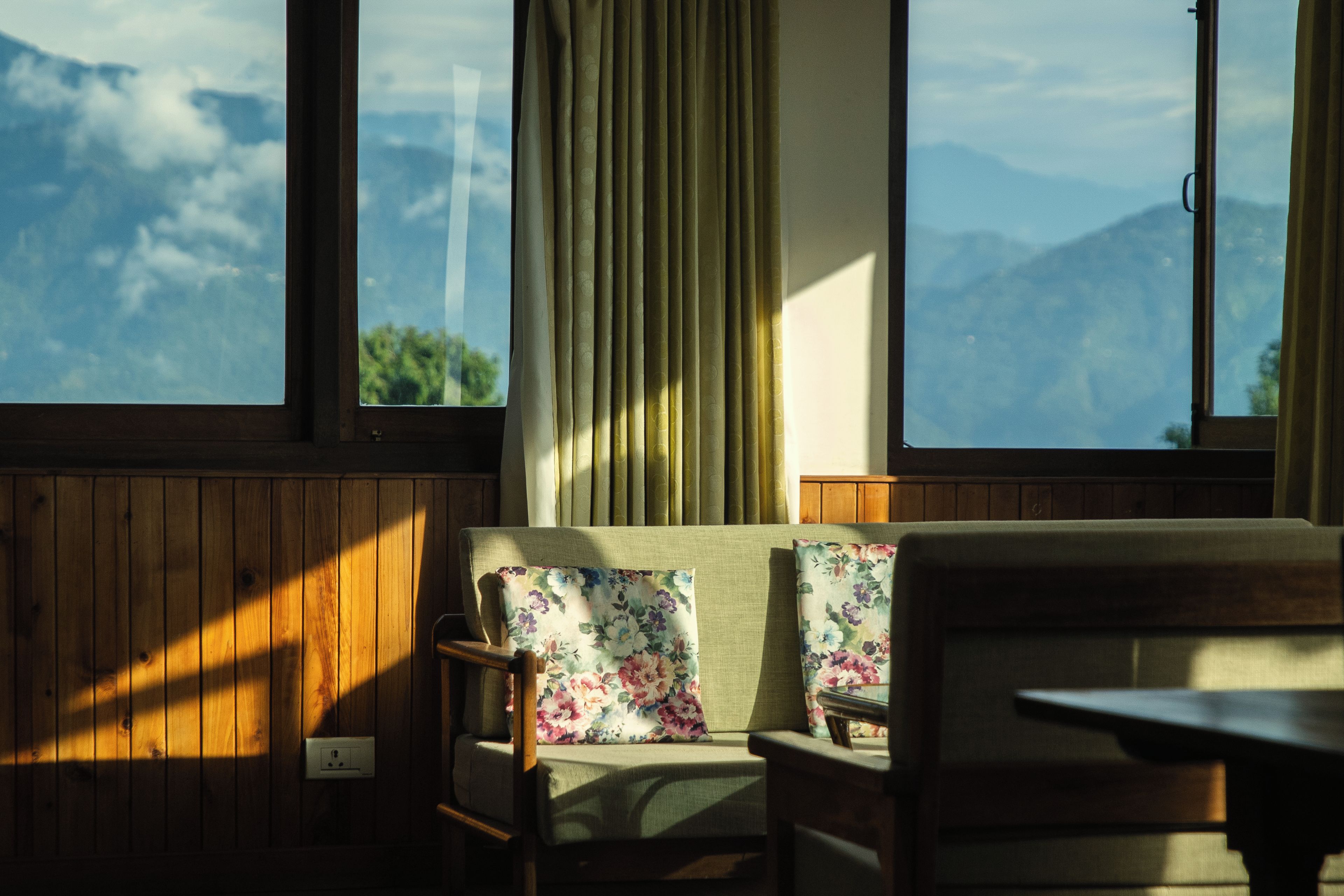 A room with a wooden paneled wall, a sofa with floral cushions, and a view of mountains through large windows.