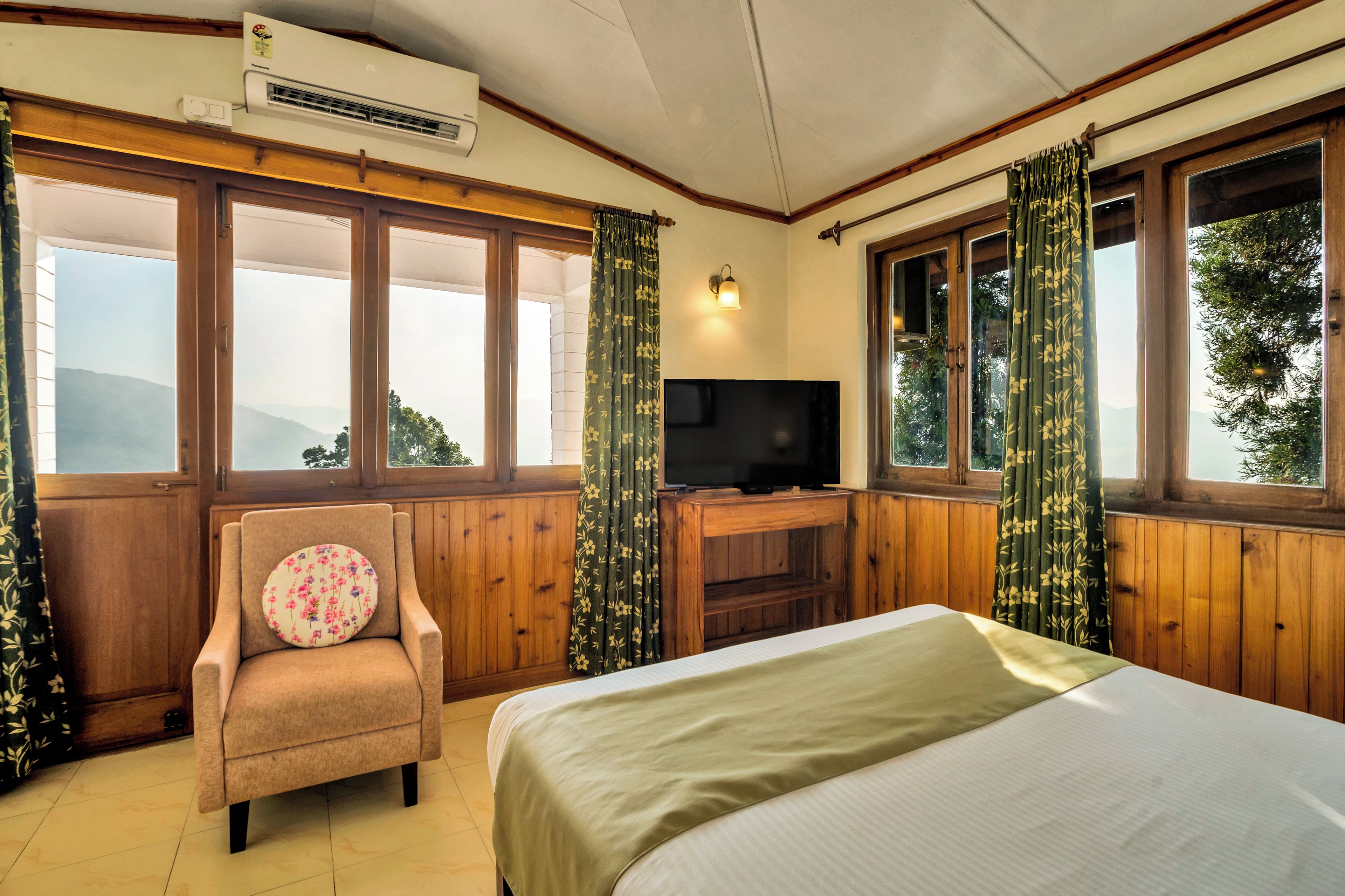 A hotel room with a bed, a chair, a television, and large windows.
