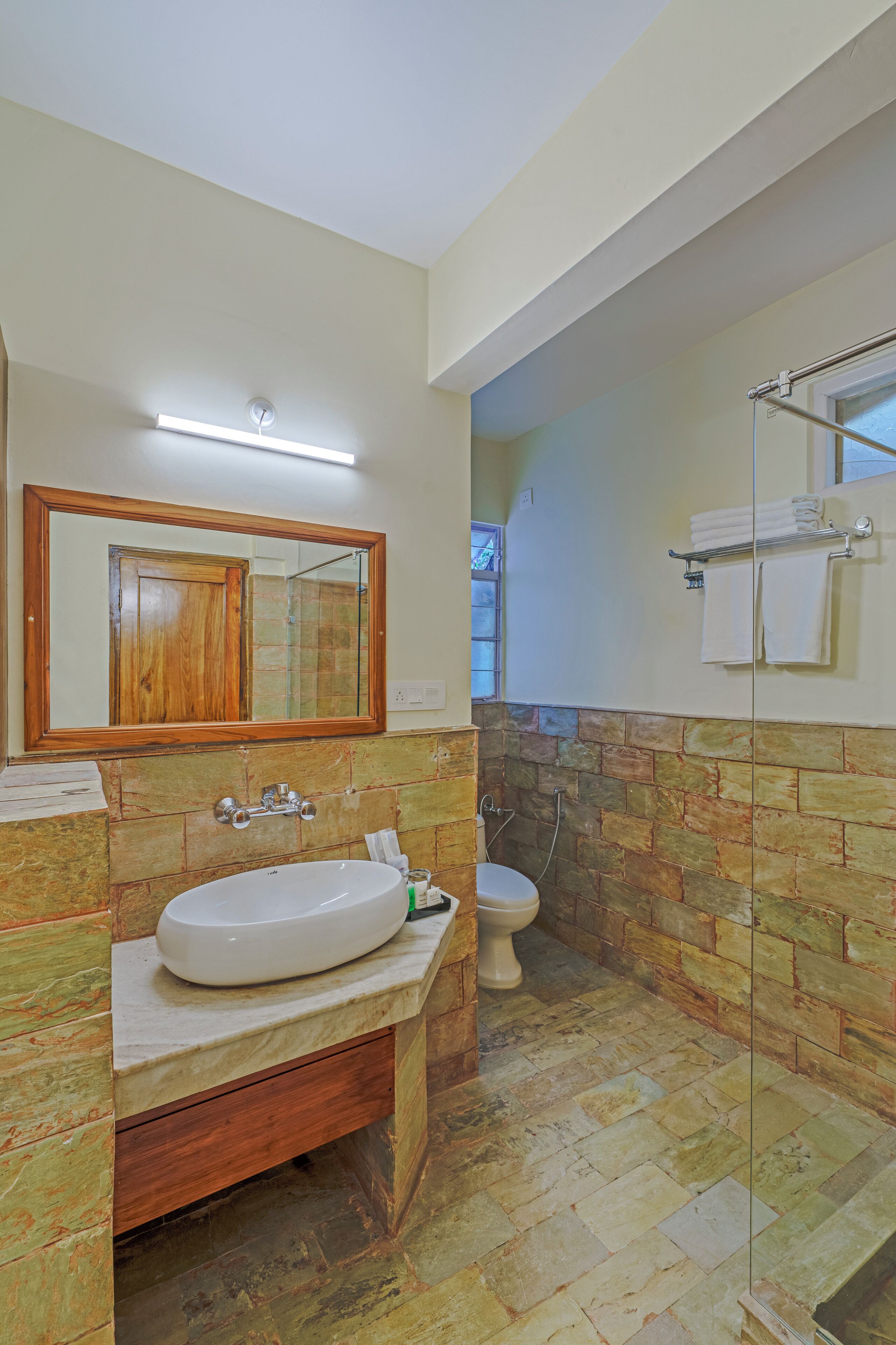 Une salle de bain avec un lavabo blanc, un miroir encadré en bois, des toilettes et un espace douche doté d’une porte en verre.