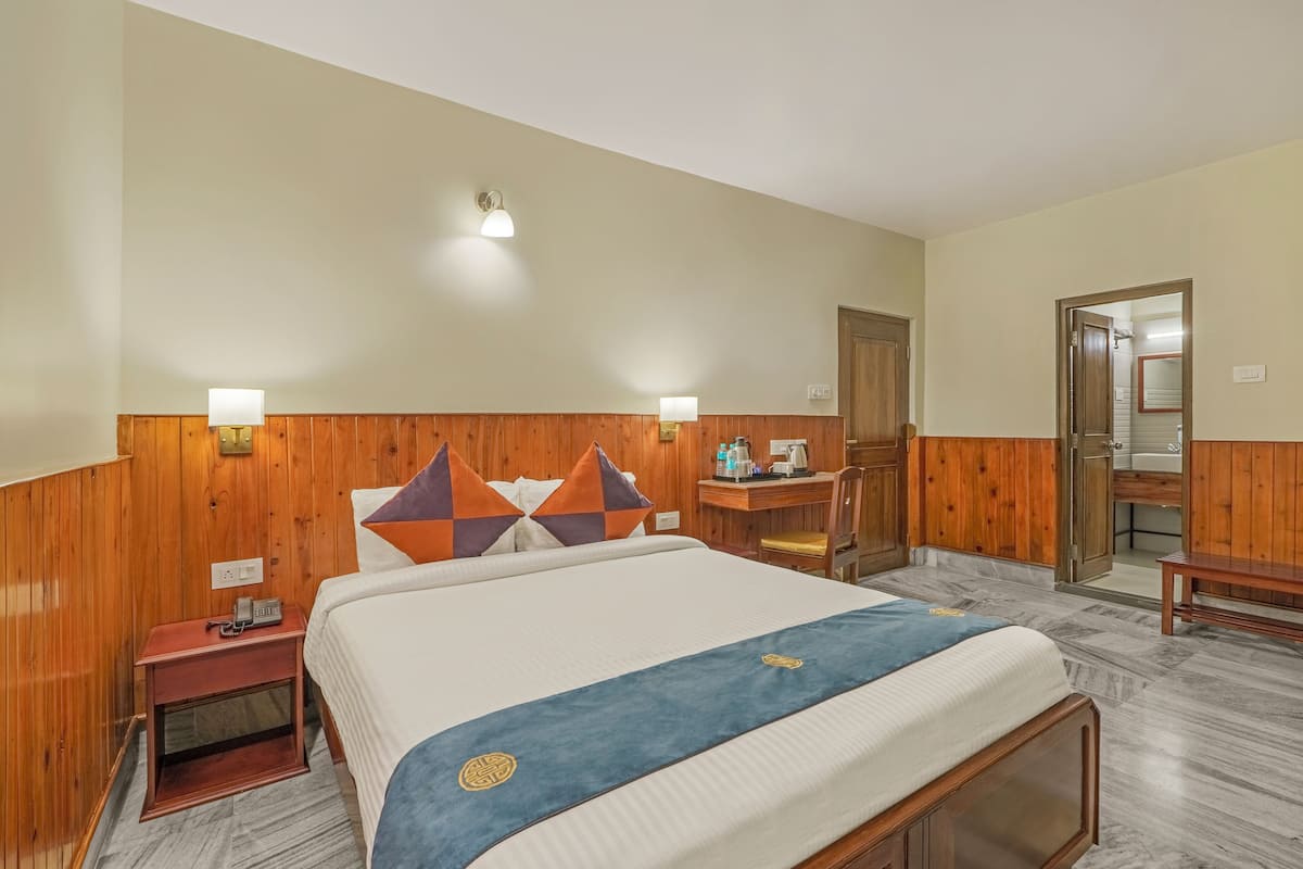 A hotel room with a wooden headboard, a bed with a blue bedspread, a nightstand with a phone, and a desk with a chair.