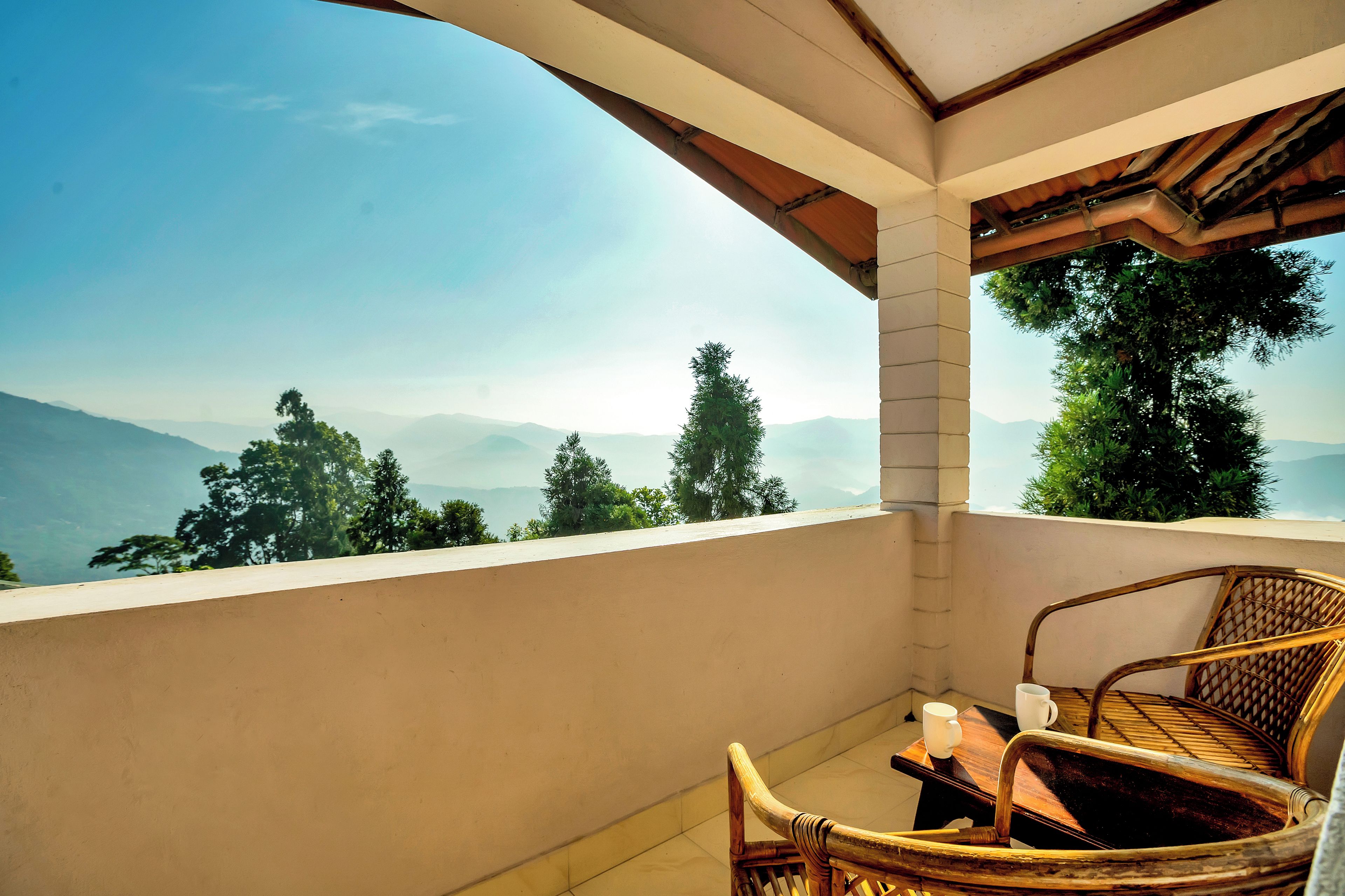A balcony with a view of mountains and trees, a wicker chair, and a table with two cups.