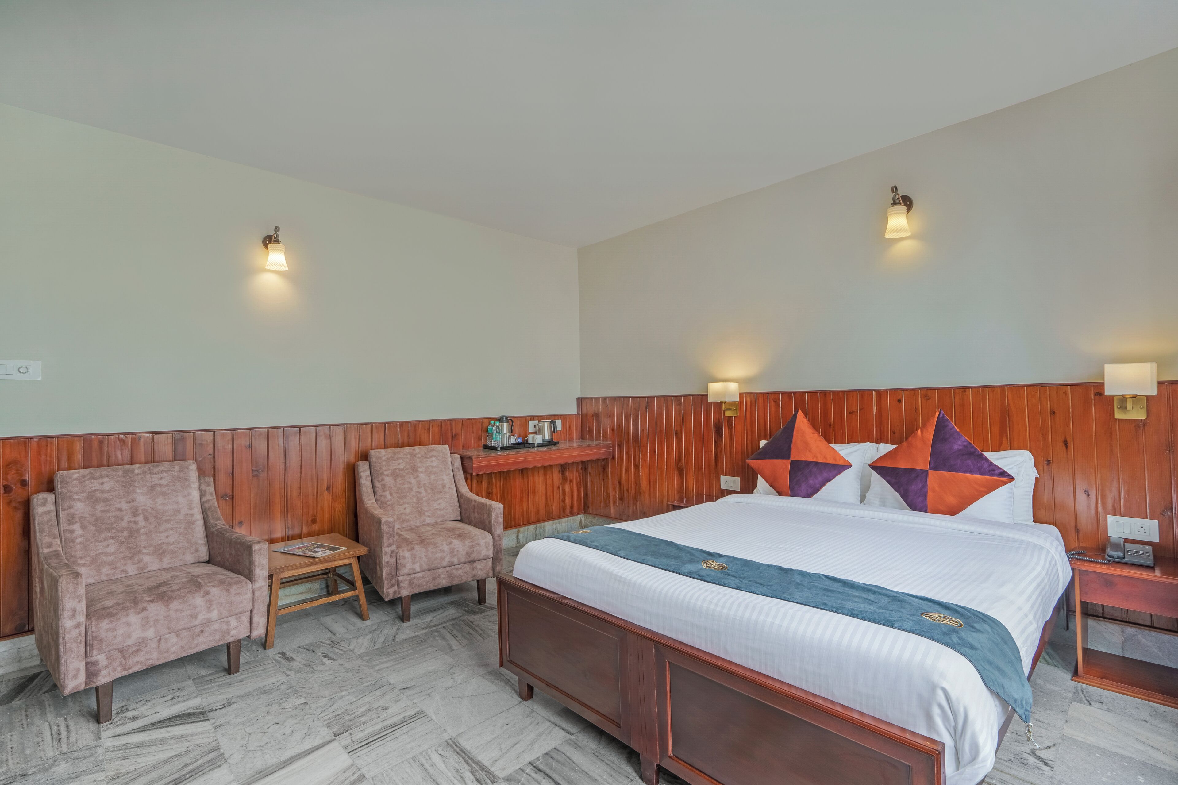 A hotel room with a large bed, two armchairs, a small table, wooden paneling, and two wall-mounted lights.