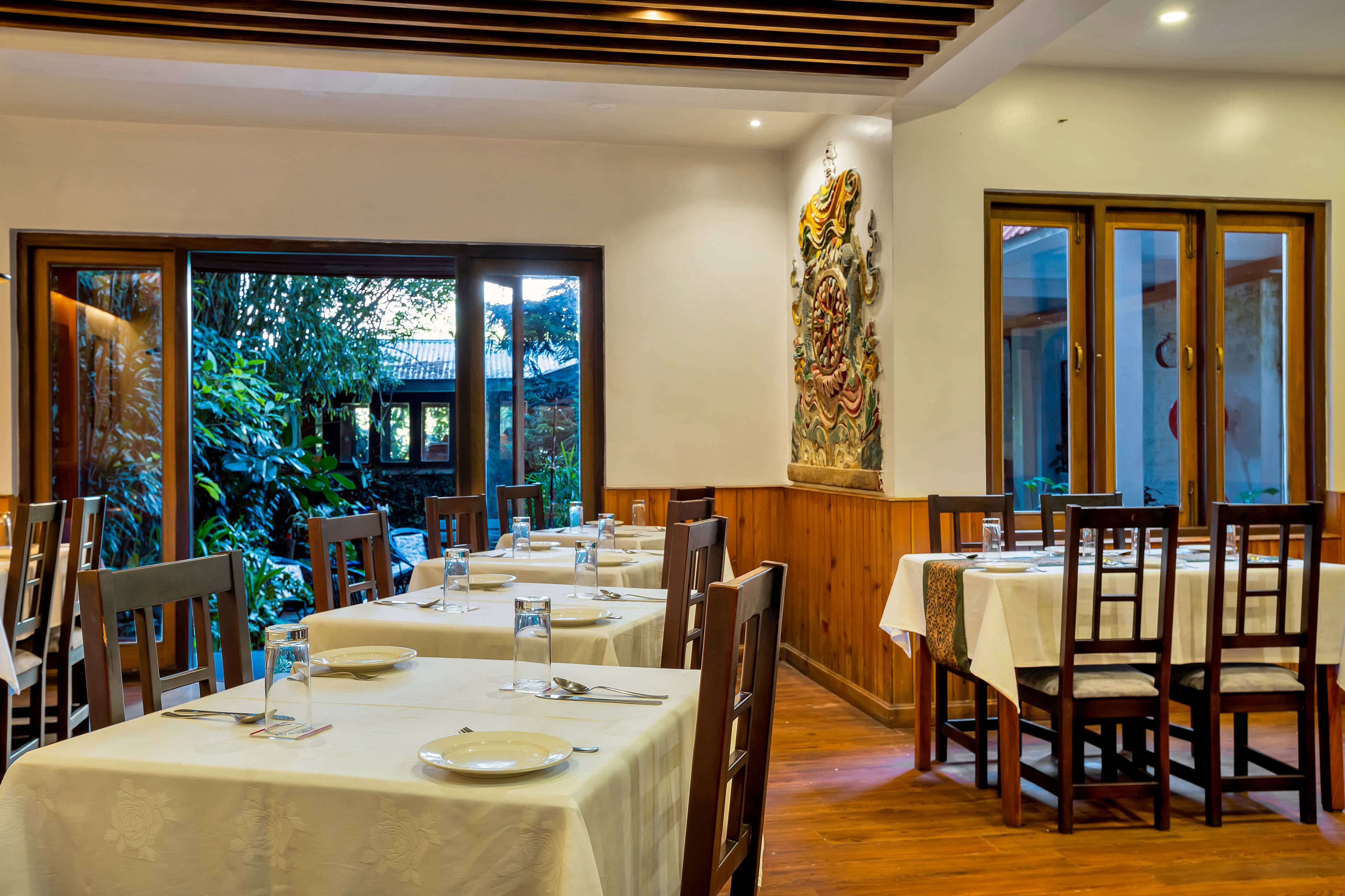 A restaurant with tables set for dining, large windows, and a view of greenery outside.