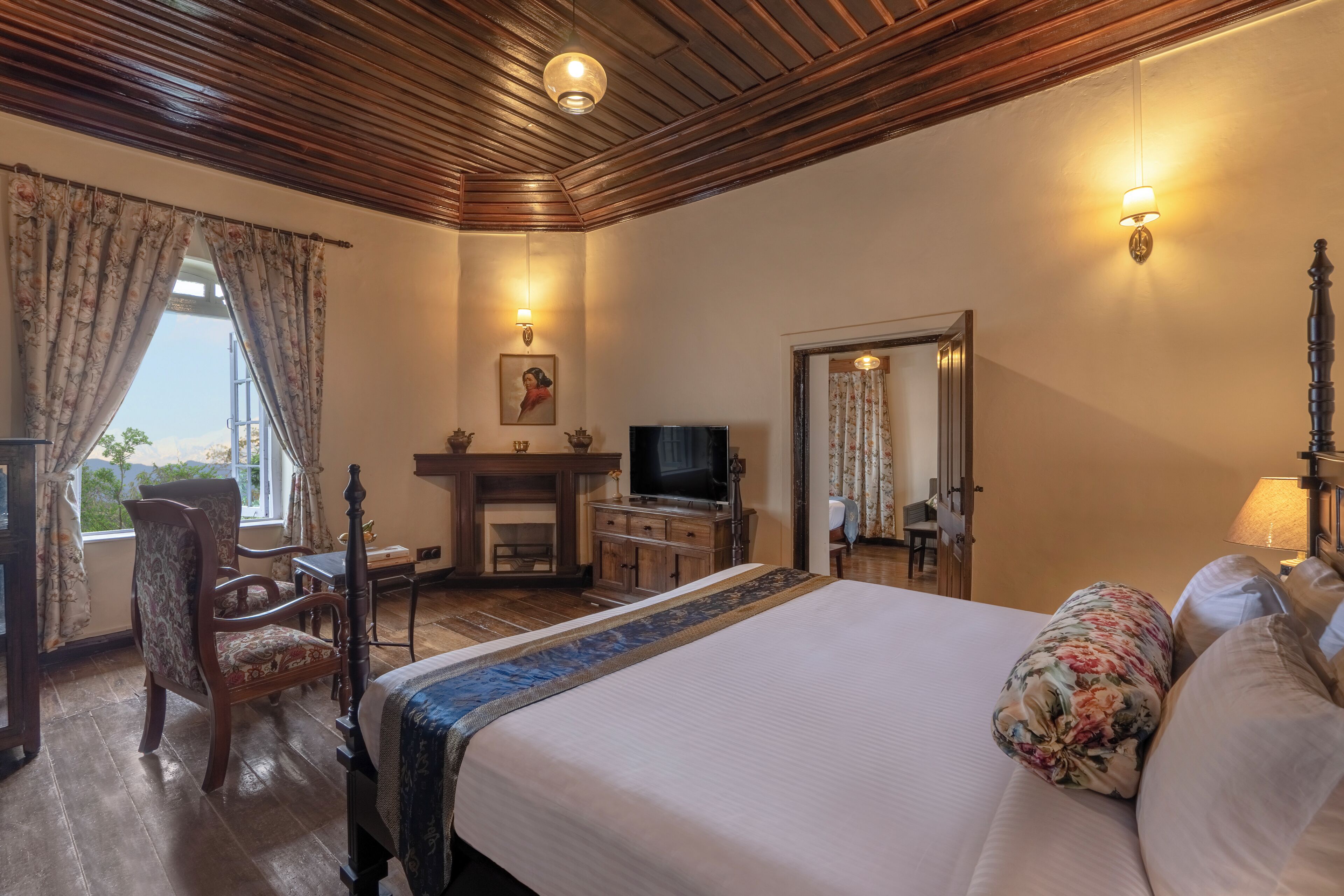 A hotel room with a large bed, wooden ceiling, a desk with a chair, a television, and a fireplace.