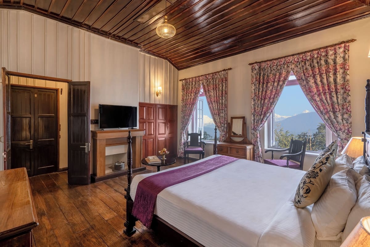 A bedroom with a wooden ceiling, a bed with a white comforter, a TV on a wooden cabinet, and a view of mountains through the window.