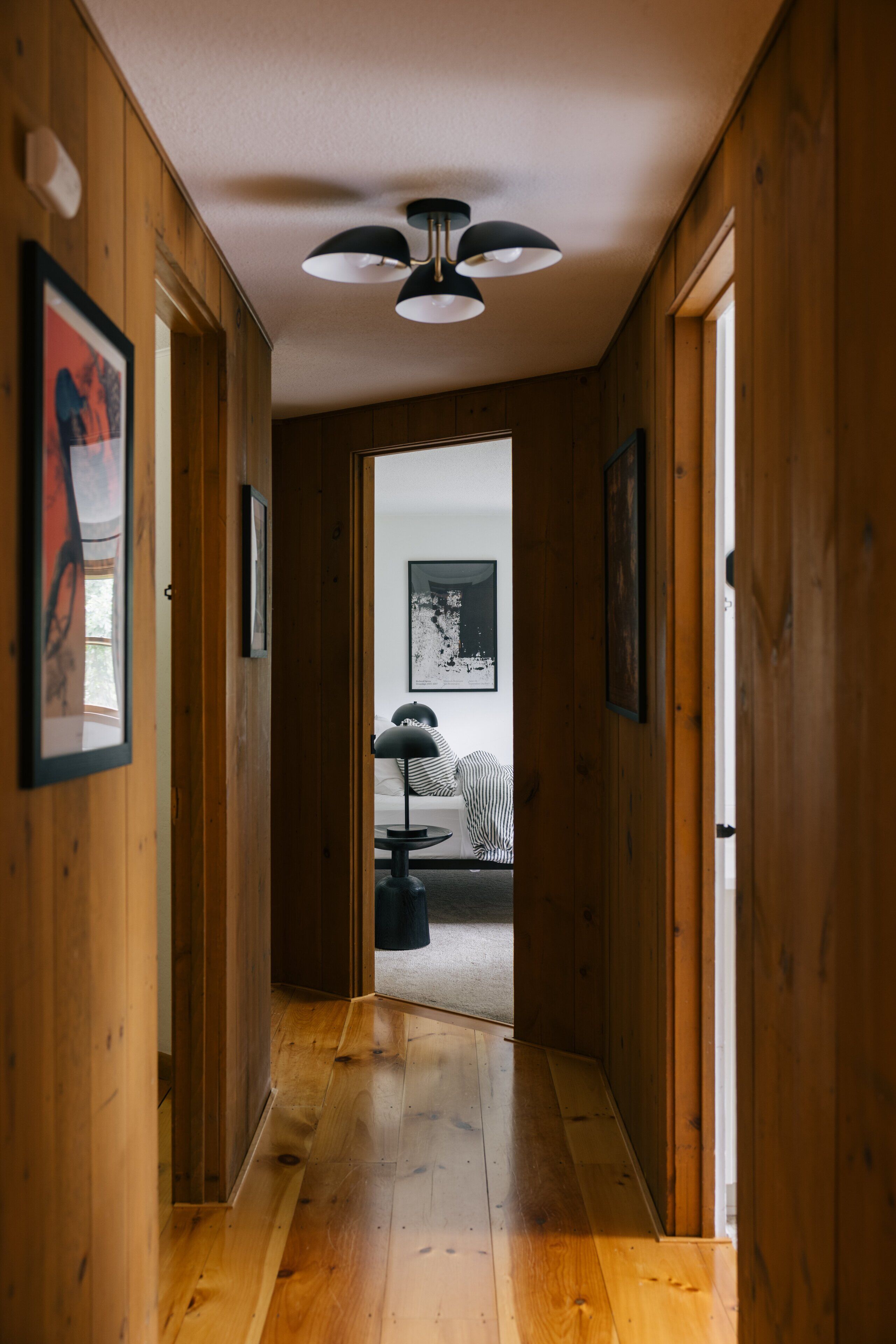 A wooden hallway leading to a bedroom with a bed, a ceiling fan, and framed pictures on the walls.
