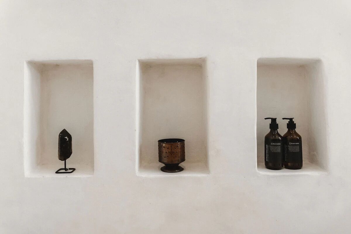 Trois niches rectangulaires sont aménagées dans un mur, et chacune contient des objets différents : un objet noir à gauche, une petite tasse en métal au centre et deux bouteilles marron à droite.
