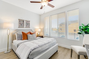 Quarto com cama, luminárias de cabeceira, ventoinha no teto e janelas amplas com persianas.