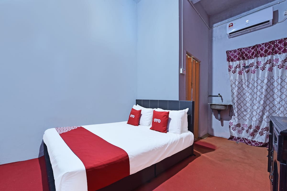 A hotel room with a bed, a red and white bedspread, two red pillows with "OYO" branding, a white air conditioning unit, a small sink, and a patterned curtain.