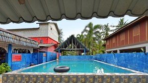 A swimming pool with stone tiles and a blue fence, surrounded by buildings and palm trees. - OYO 90664 Rabi Hotel (Bachok)