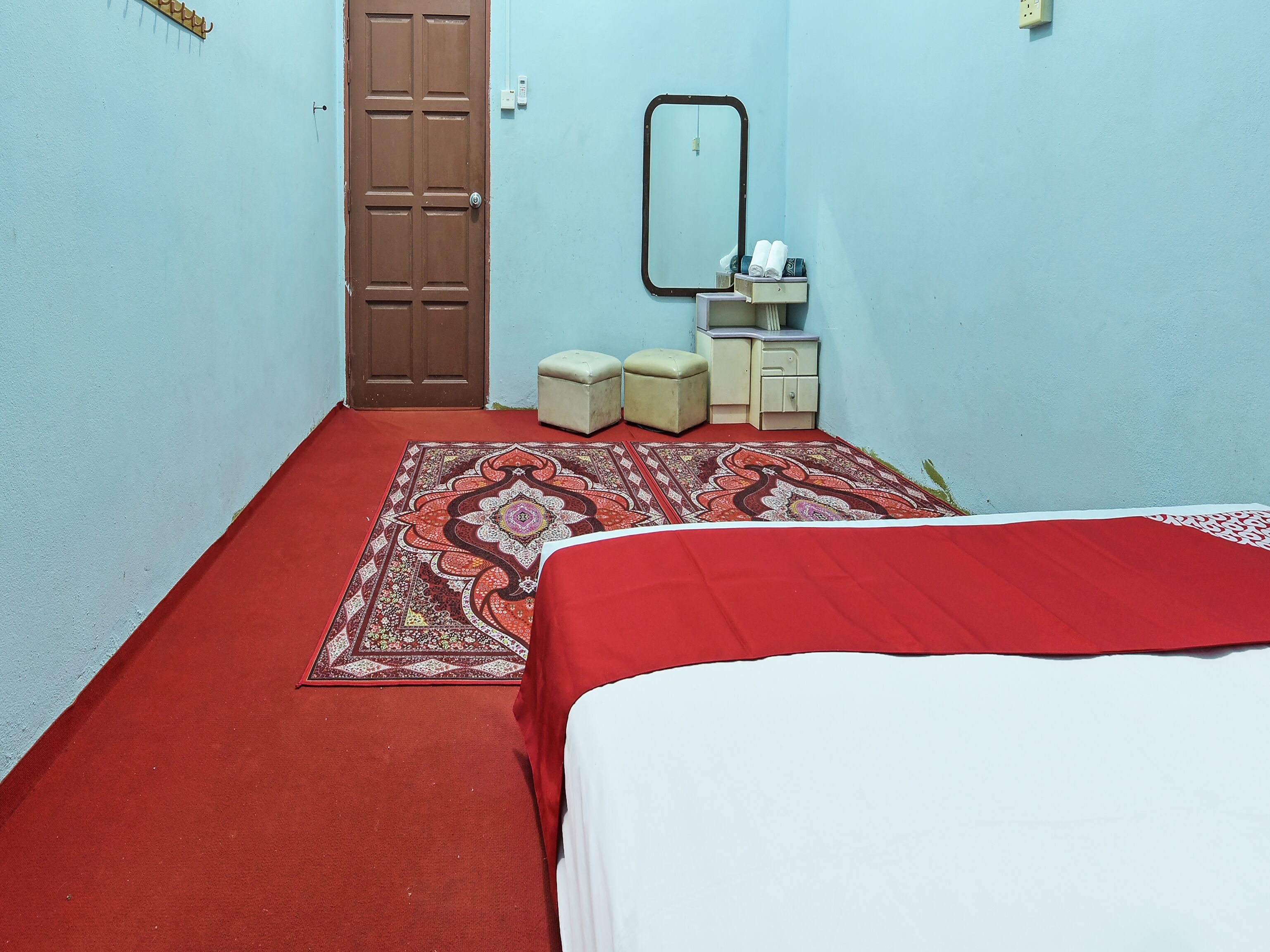 A bedroom with a red carpet, a bed with a white cover, a small bedside table, a mirror, and a wardrobe.