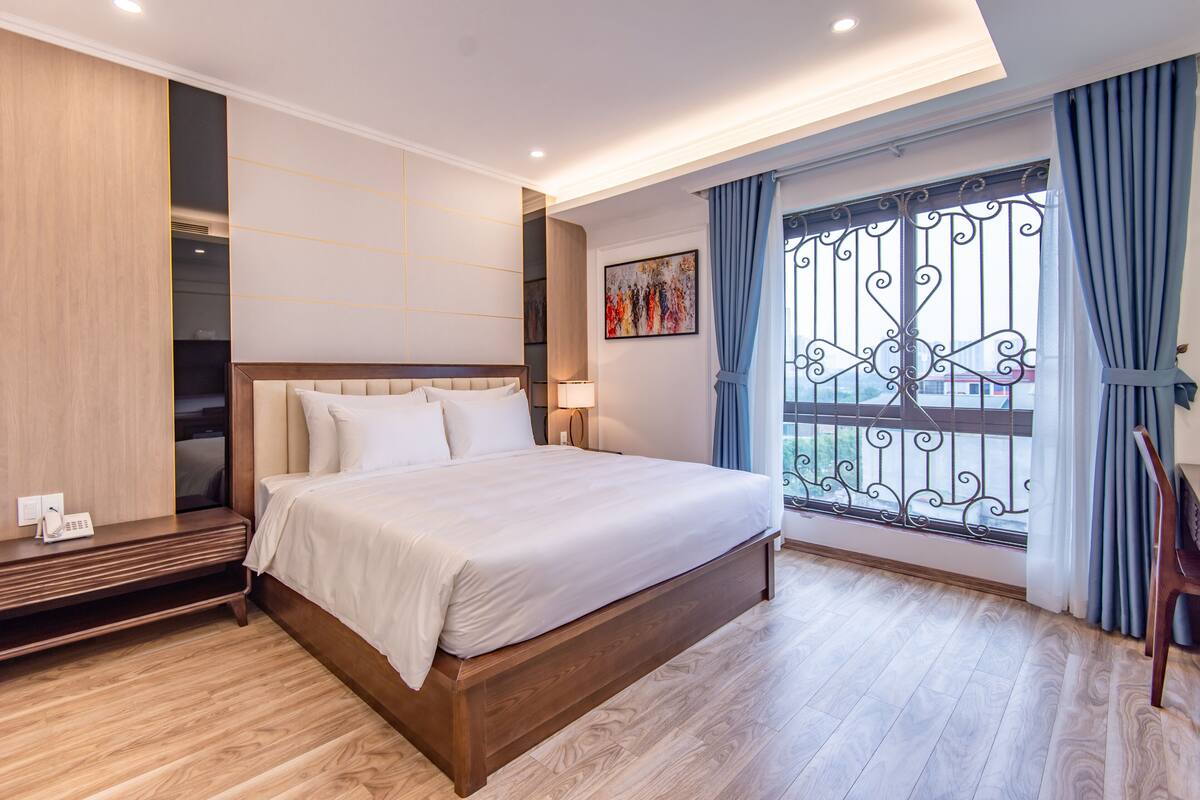 A modern hotel room with a large bed, wooden flooring, a bedside table with a lamp, and a window with decorative iron bars.