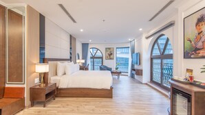 Quarto de hotel espaçoso com uma cama grande, área de estar com poltrona e mesa, e janelas amplas com vista.