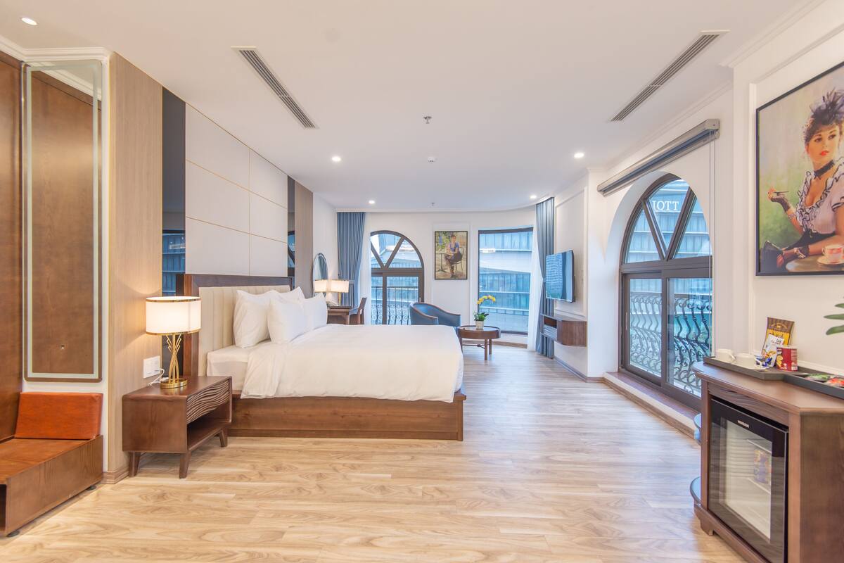 A spacious hotel room with a large bed, a sitting area with a chair and table, and large windows offering a view.