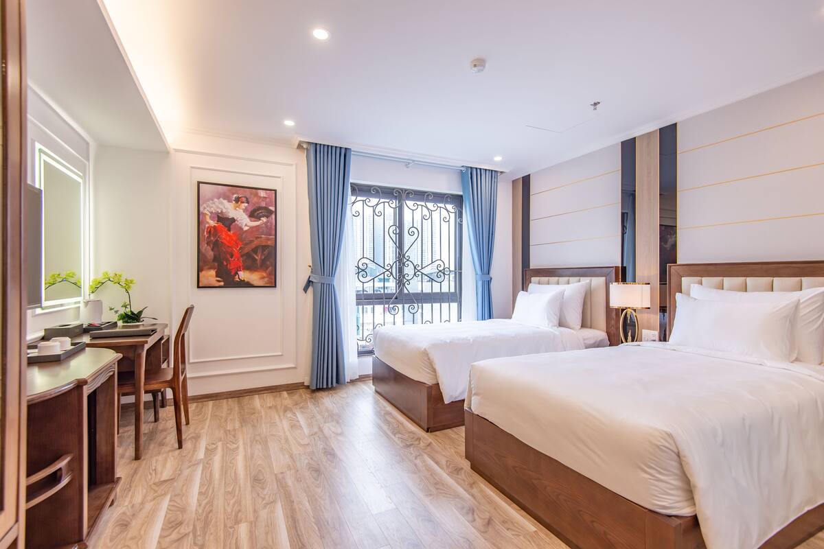 A hotel room with a wooden floor, two beds, a desk with a chair, a large window with curtains, and a painting on the wall.