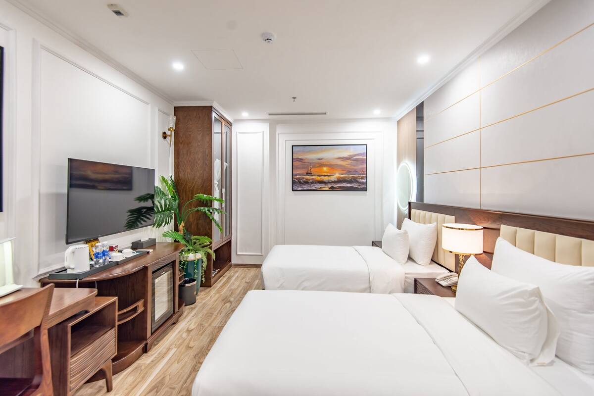 A hotel room with two beds, a desk, a TV, a painting, and a plant.