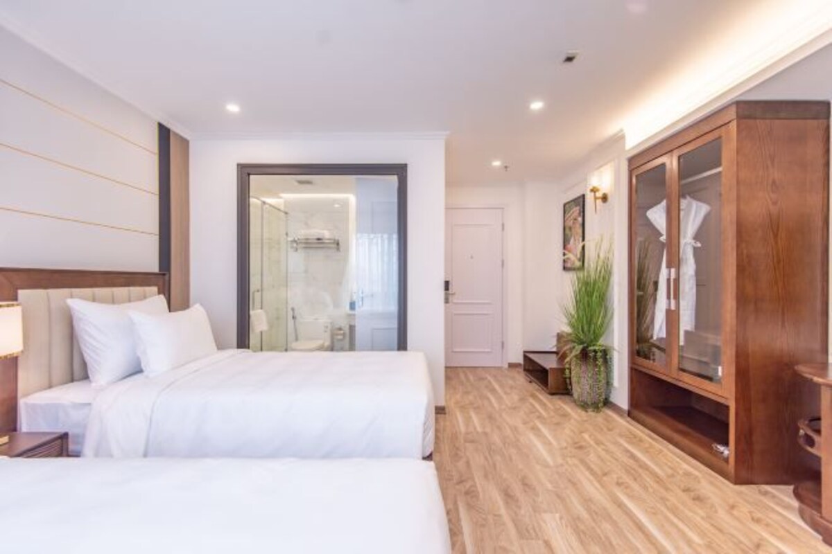 A modern hotel room with two beds, a wooden wardrobe, a potted plant, and a glass shower enclosure.