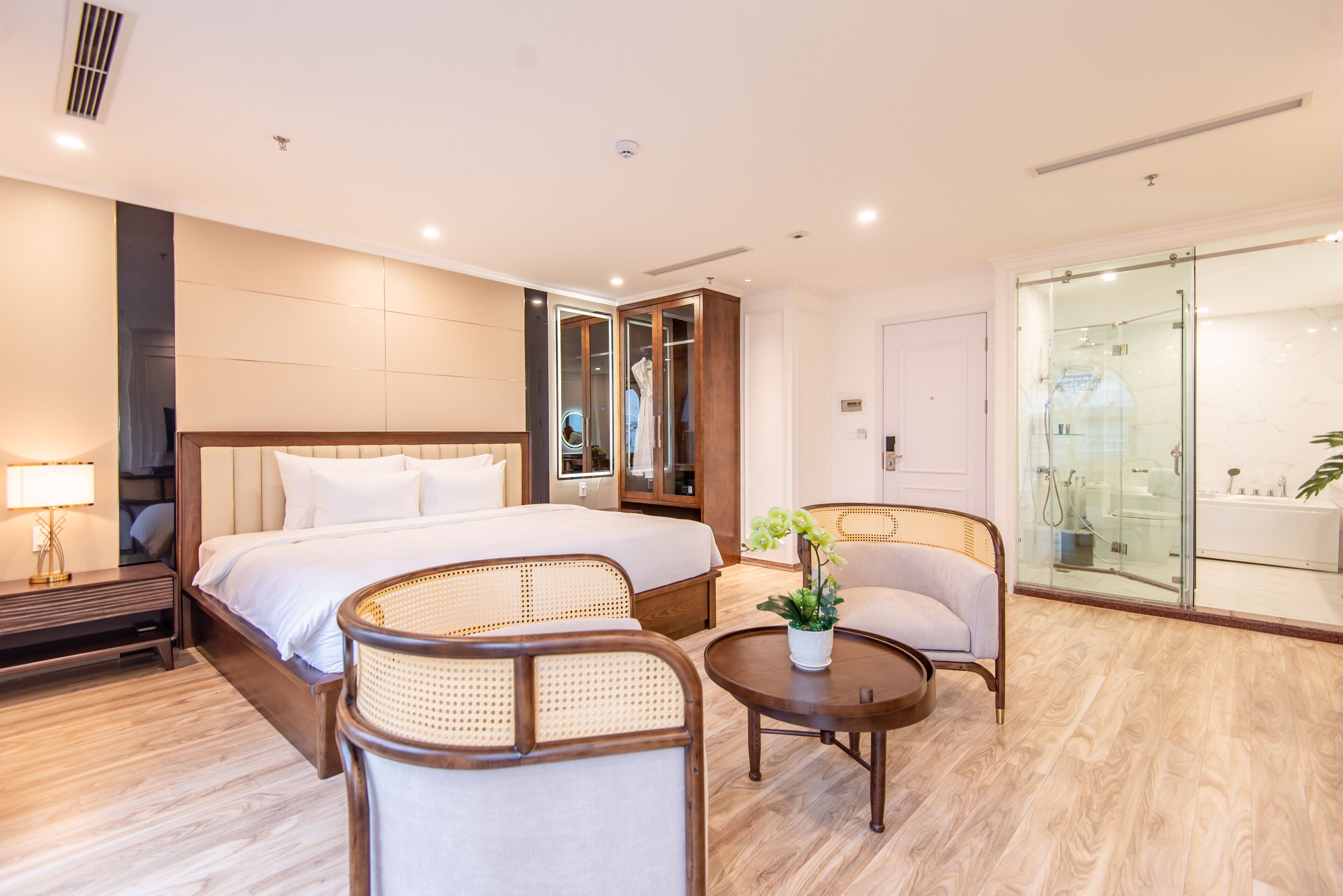 Une chambre d’hôtel moderne dotée d’un grand lit, d’un coin salon avec un fauteuil et une petite table, d’une douche en verre et d’une armoire avec un miroir.