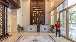 Um saguão de hotel com uma mesa de recepção, uma porta de madeira decorativa e grandes janelas que oferecem vista para o exterior.