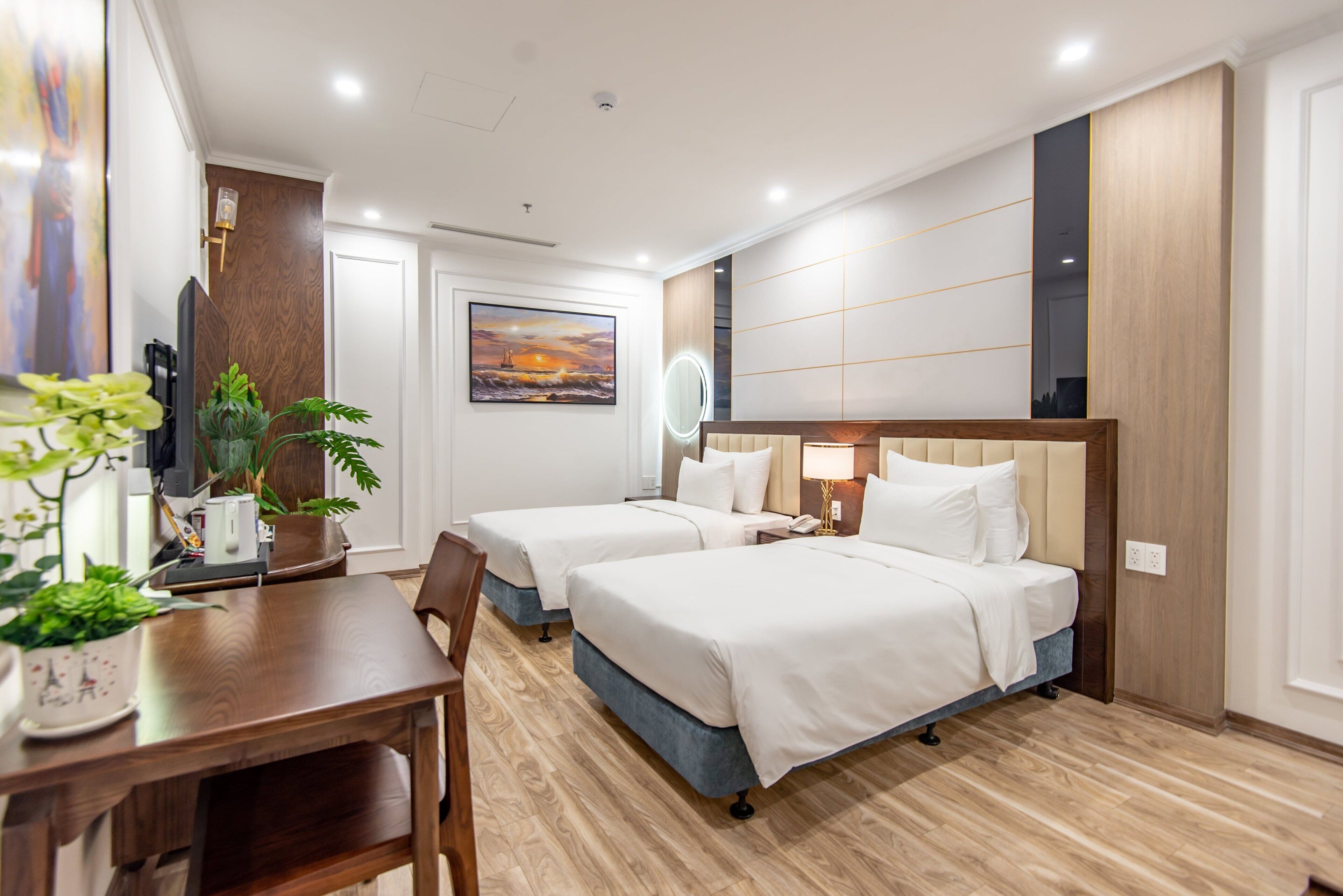 A hotel room with two beds, a wooden floor, a desk with a chair, a TV, and a painting on the wall.