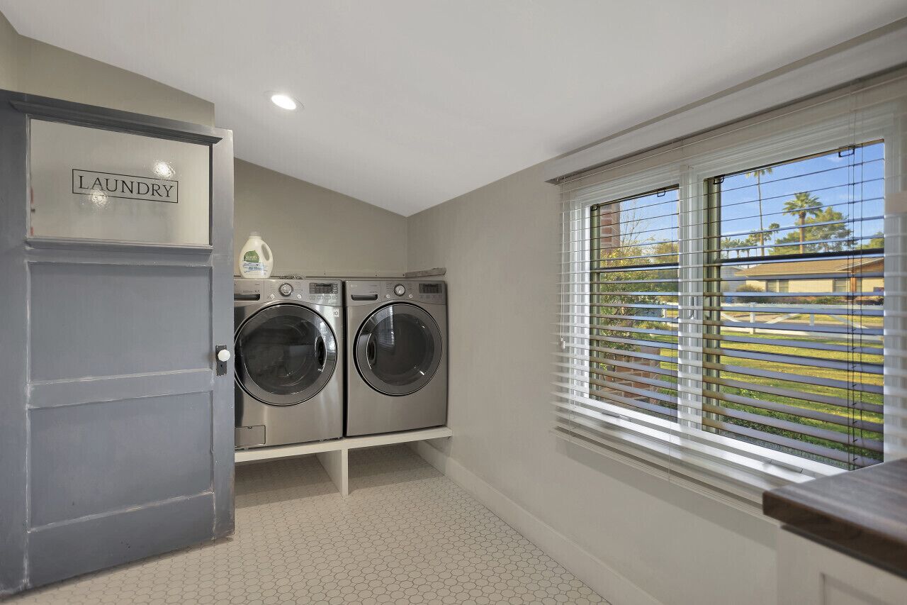 A laundry room with two front-loading washing machines, a built-in cabinet labeled 'laundry', and a window with blinds overlooking a residential area.