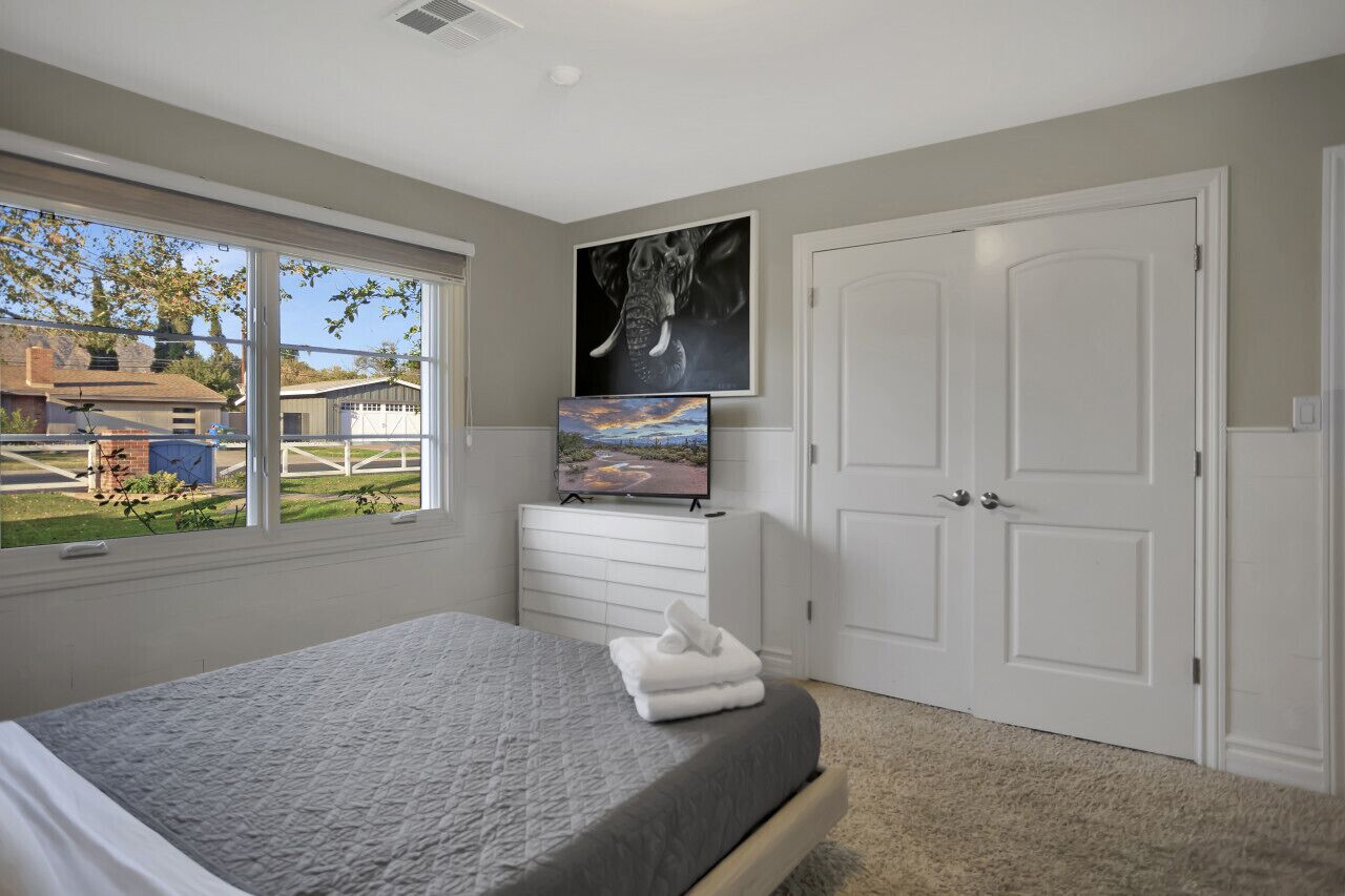 A bedroom with a bed, a dresser, a TV, and a large window.