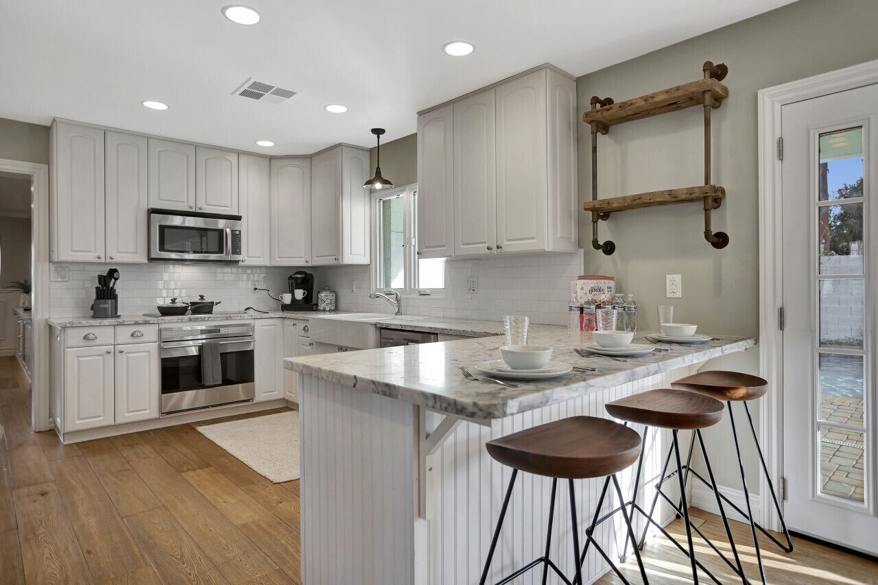 Una cocina moderna con armarios blancos, una isla central con taburetes de bar, un estante de madera colgante y un ventanal que proporciona luz natural.