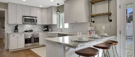 Una cocina moderna con armarios blancos, una isla central con taburetes de bar, un estante de madera colgante y un ventanal que proporciona luz natural.