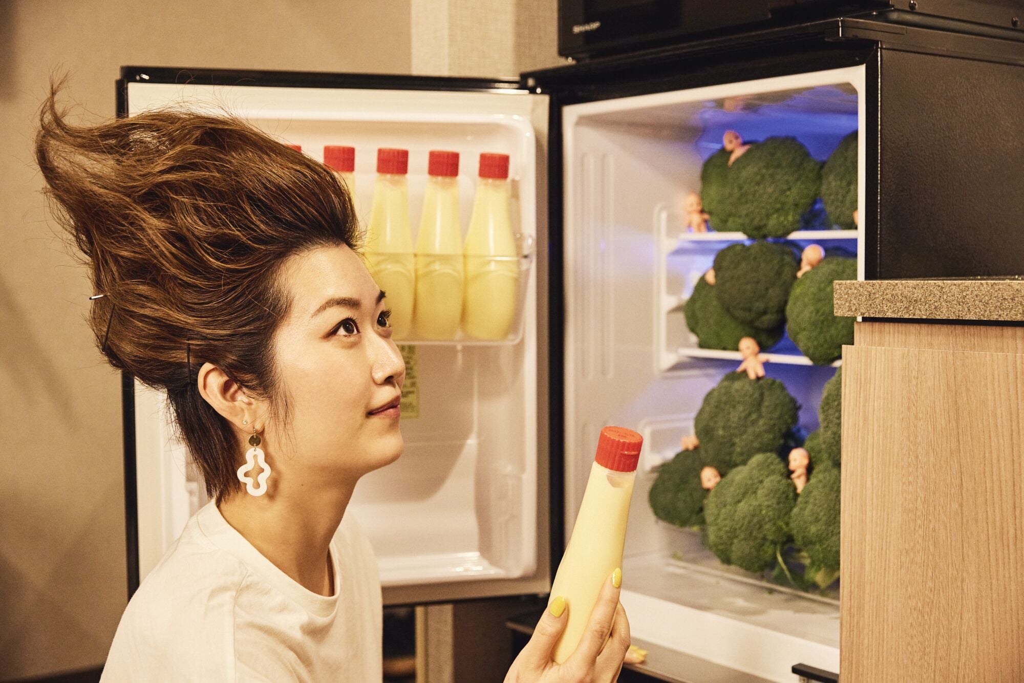 A person holding a bottle in front of an open refrigerator with bottles and broccoli inside.