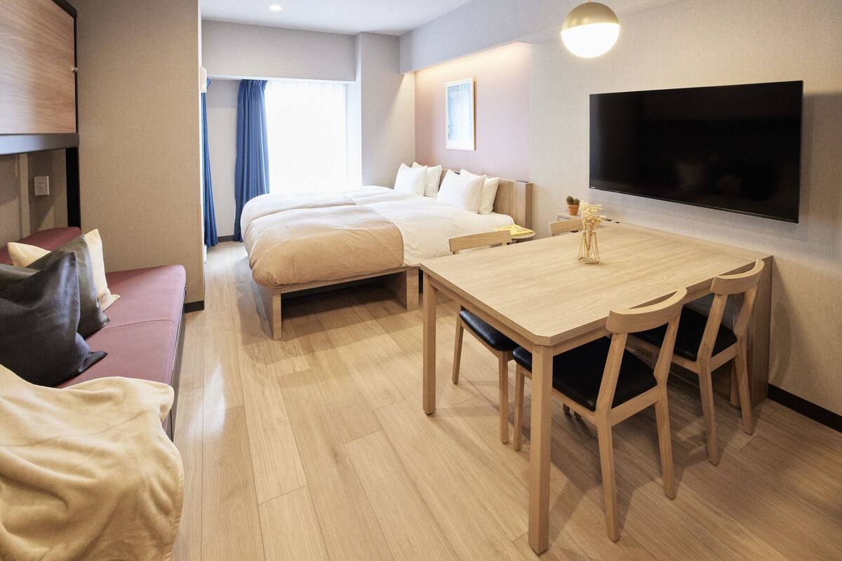 A hotel room with a bed, a desk with chairs, a TV, and a window with curtains.