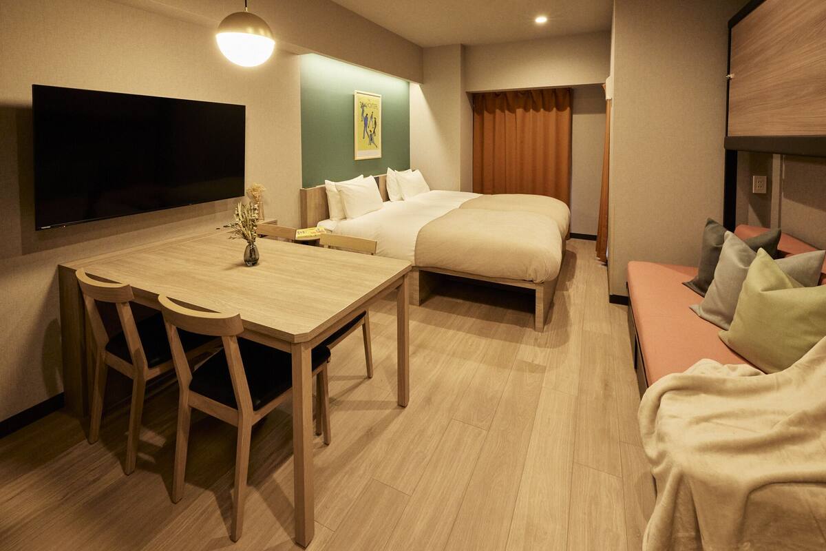 A hotel room with a bed, a desk with chairs, a TV, and a small table with a vase.