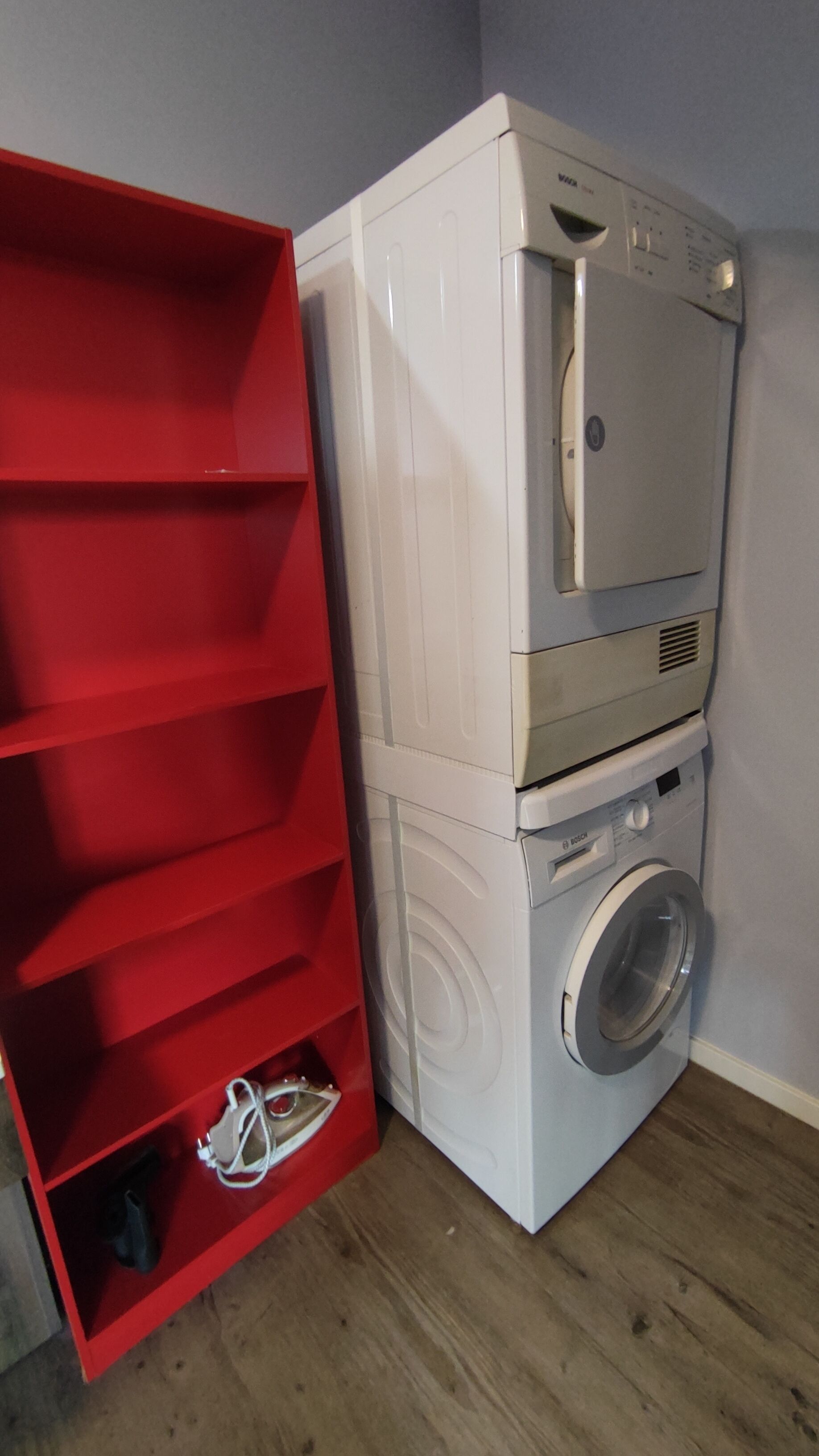 Een kleine wasruimte met een gestapelde wasmachine en droger, een rode bergkast met planken, en een houten vloer.