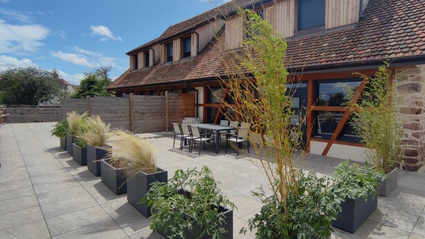 Un bâtiment moderne doté d’une façade en pierre, d’un toit en tuiles et d’un patio aménagé avec des espaces de détente en plein air et des jardinières.