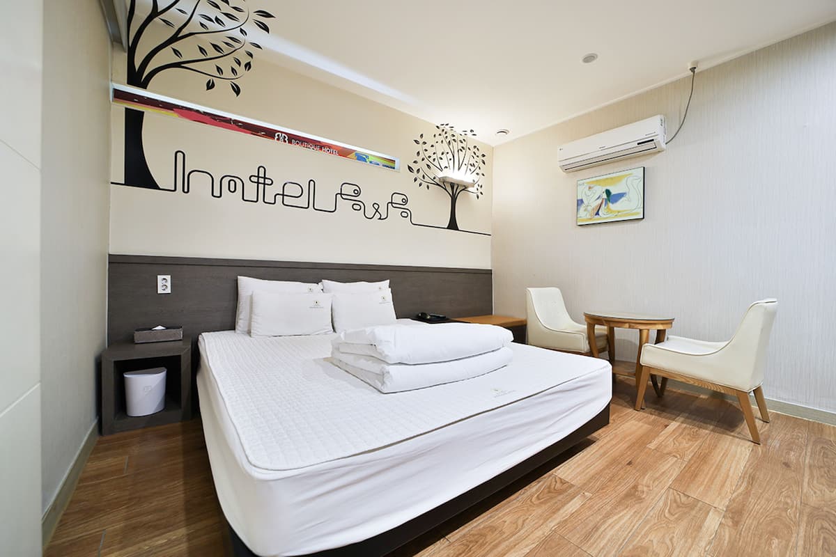 A hotel room with a large bed, a nightstand, a chair, a small table, and a wall-mounted air conditioner.