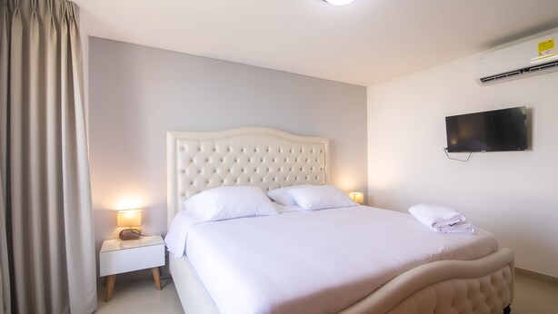 Een net opgemaakt bed met een gestoffeerd hoofdbord, een bedlamp en een wandgemonteerde flatscreen-tv.