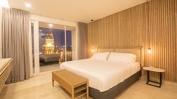 A modern hotel room with a large bed, a bench, a small table, and a view of a cityscape with a clock tower. - 7 Bedroom in Clock Tower Plaza with Private Pool (Cartagena)