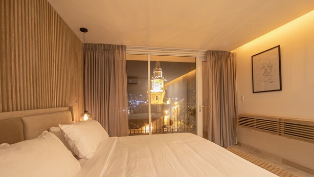 A hotel room with a large bed, a view of a cityscape with a clock tower, and a framed picture on the wall. - 16 Bedroom in Clock Tower Plaza with Private Pool (Cartagena)
