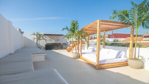 Una terraza en la azotea con una cama con dosel de madera, sillones blancos para lounge y plantas en macetas.
