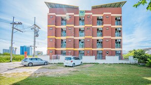 A multi-story building with a red and yellow facade, air conditioning units, and a parking area with two cars. - Supatranai Home Cha-am (Cha-am)