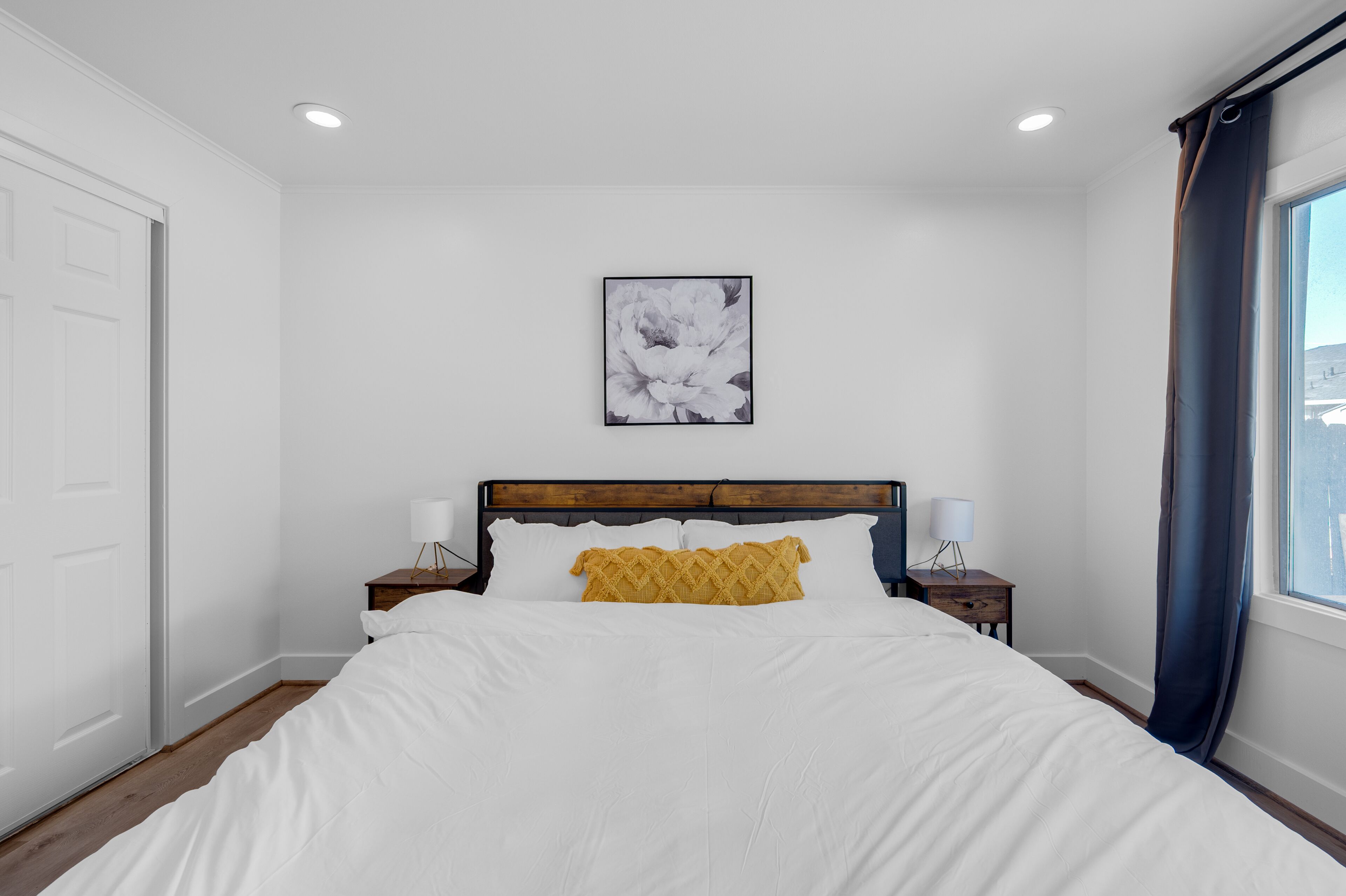 A bedroom with a bed, a nightstand, a wall-mounted picture, and a window with curtains.