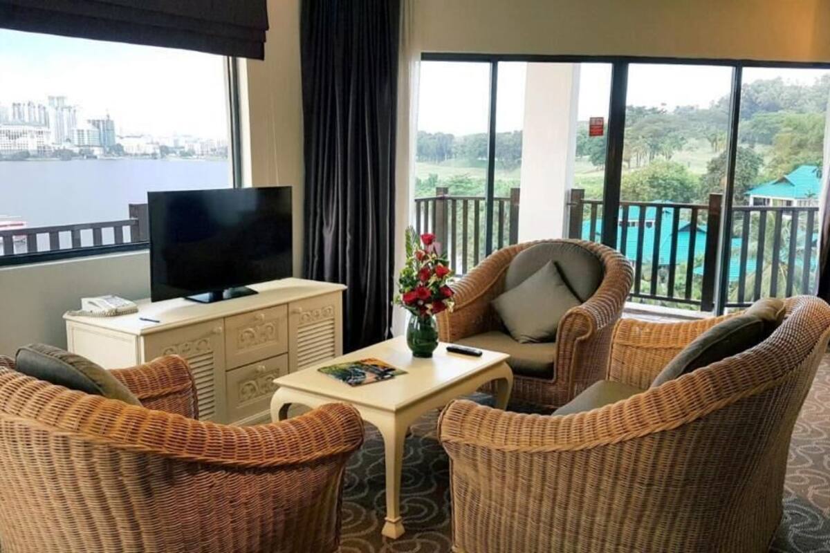 A hotel room with wicker furniture, a television, a small table with a vase of flowers, and large windows offering views of a city and a pool.