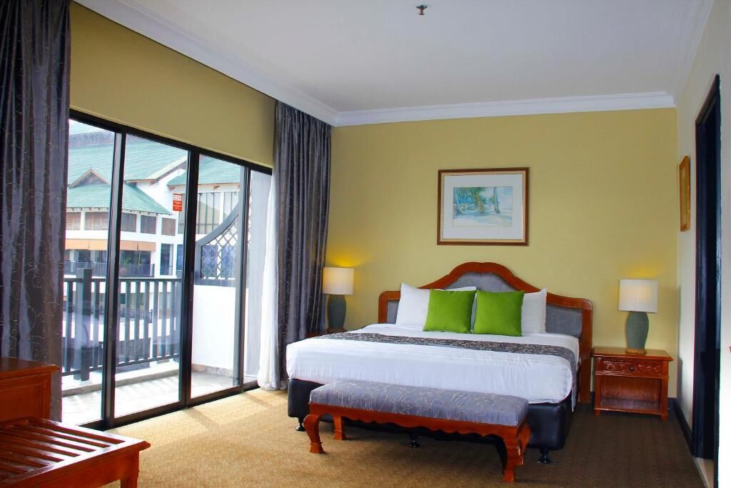 A hotel room with a large bed, a wooden bench, a nightstand, and a view of the outside balcony.
