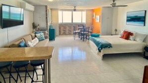Un'ampia zona giorno con divano, tavolo da pranzo con sedie, letto con cuscini, TV a parete e vista sull'esterno attraverso una grande finestra.