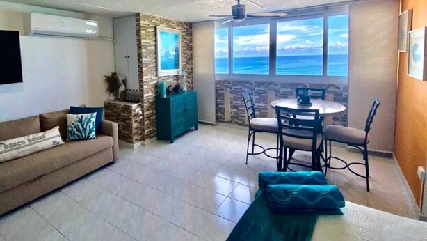 Un soggiorno con divano, un tavolo da pranzo con sedie, una ventola a soffitto e vista sull'oceano attraverso ampie finestre.