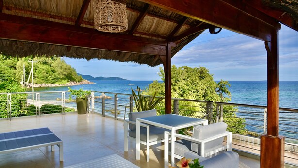 A covered outdoor seating area with a view of the sea, white furniture, and a thatched roof. - Sea Splash & La Petite Maison (Praslin Island)
