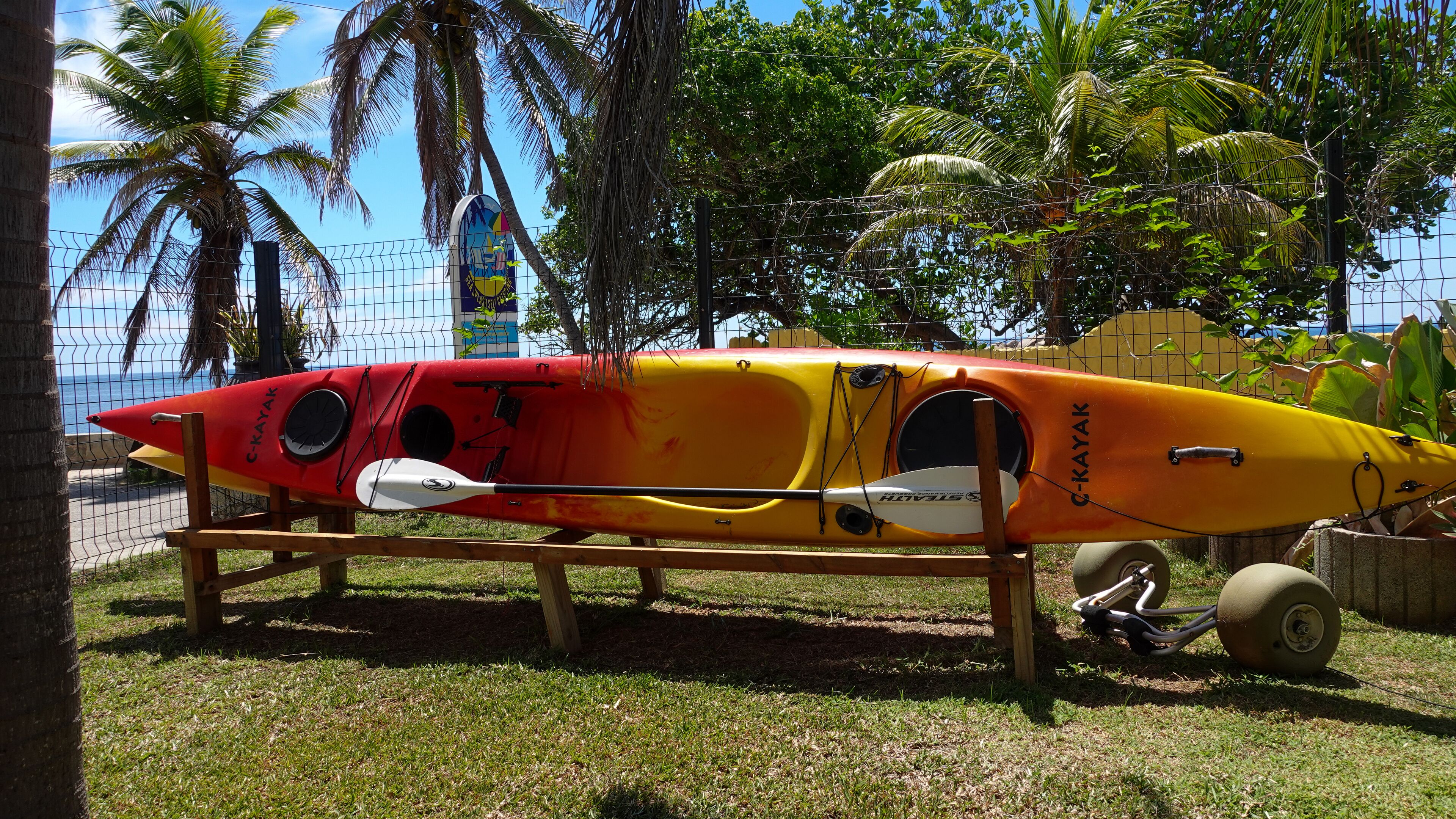 A kayak with a red and yellow design, placed on a wooden stand in a grassy area with palm trees and a fence in the background.