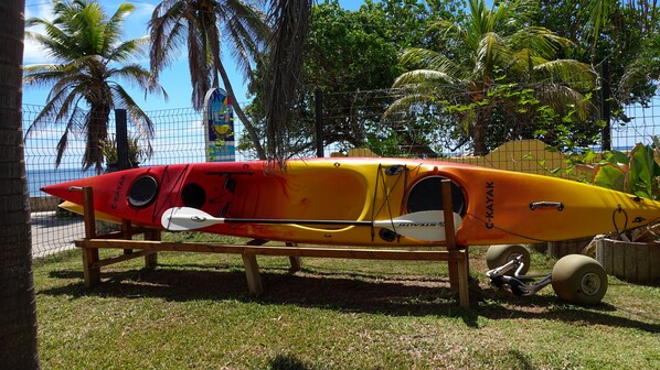 A kayak with a red and yellow design, placed on a wooden stand in a grassy area with palm trees and a fence in the background. - Sea Splash & La Petite Maison (Praslin Island)